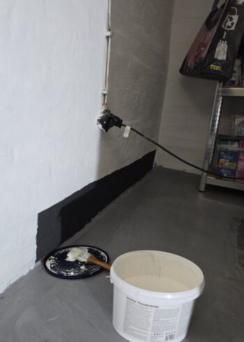 Garage corner with white wall and black baseboard being painted; paint bucket and tray with brush on the floor; cord plugged into wall near a shelf of supplies.