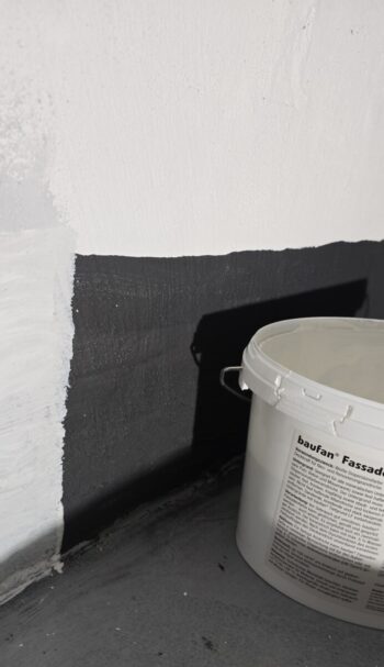 White paint bucket with handle sits next to a two-tone wall (white upper, dark gray lower).